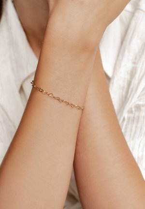 Gold chain bracelet on a wrist, featuring linked oval shapes with a polished finish, resting against a light-colored textured fabric background.