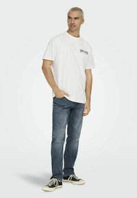 Maglietta di cotone bianca con un logo stampato nero sul lato sinistro del petto, abbinata a jeans in denim blu e sneakers nere con suole bianche.