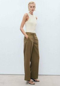 Vit ribbad tanktopp i kombination med högmidjade, vida bruna byxor. Modellen står med händerna i fickorna och bär svarta sandaler med remmar.