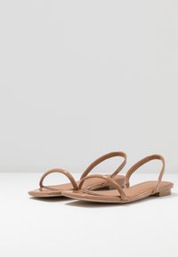 Vaaleanruskeat litteät sandaalit, joissa on kaksi hoikkaa, läpinäkyvää hihnaa ja sileä, kiiltävä pinta. Sisältää minimalistin muotoilun ja litteän pohjan.