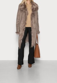 Persona vistiendo un abrigo de imitación de piel en color topo con borde de piel sintética, suéter beige, pantalones negros, botines marrones de tacón, y sosteniendo un bolso de mano de piel marrón.