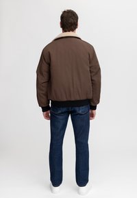 Hombre con una chaqueta bomber marrón con cuello de piel clara, vaqueros azul oscuro y zapatillas blancas, de pie de espaldas sobre un fondo liso.