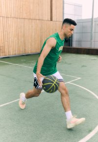 A man dribbles a basketball wearing a green sleeveless shirt and black shorts with a white accent, on a green court with wooden walls.