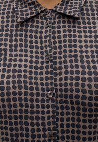 Chemise avec des pois noirs irréguliers sur tissu beige, dotée de cinq boutons noirs et d’un col pointu.