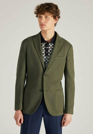 Blazer vert olive avec revers en pointe, fermeture à un bouton, deux poches avant et tissu texturé. Porté sur une chemise polo à motif.