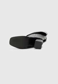 Black heeled shoe with a smooth leather upper, square toe, and chunky heel. The sole features a textured grip pattern for traction.