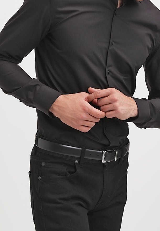 Homme portant une chemise noire boutonnée et un pantalon noir, en train d'attacher une ceinture en cuir noire à la taille.