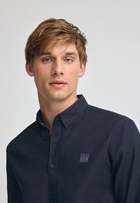 Camicia blu navy con bottoni, in tessuto testurizzato, maniche lunghe e una piccola toppa con logo sul petto. Design del collo classico.
