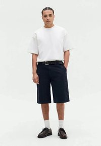 Hvit oversized t-skjorte, mørke stripete shorts til kneet, svart belte, hvite sokker og svarte sko. Enkel bakgrunn.