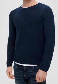 Mies, joka pukeutuu teksturoituun tummansiniseen pitkähiharaiseen neuleeseen valkoisen aluspaidan päällä tummien farkkujen kanssa, seisoo vaaleaa taustaa vasten.