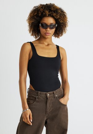 Žena s kučeravými vlasmi, oblečená v čiernom tielku bez rukávov, hnedých nohaviciach s rukou v vrecku, tmavými okuliarmi s obručou a strieborným náramkom.