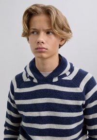 Teini-poika, jolla on keskipitkä vaaleanblondi hius, pukeutuneena tummansiniseen ja valkoiseen raidoitettuun huppariin, katsoo sivulle yksinkertaista taustaa vasten.