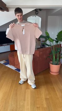 Light pink short-sleeve t-shirt displayed, featuring a round neckline and tag attached, held by a person in casual beige pants and sneakers.