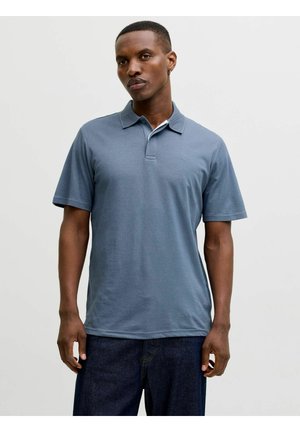 Homme portant un polo à manches courtes bleu et un jean foncé, debout devant un fond clair uni.