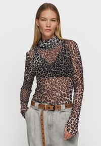 Durchsichtige Bluse mit langen Ärmeln im Leopardenmuster in Brauntönen und Schwarz. Mit einem hohen Kragen und figurbetontem Schnitt, kombiniert mit hochtaillierten, hellgrauen Hosen.