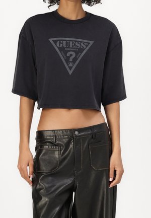 Beskåret billede af person iført sort cropped Guess t-shirt og sort læderbukser med højtaljet, stående med arme afslappet ned langs siden.