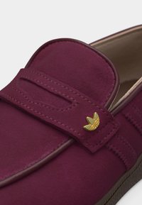 Close-up van een bordeauxrode suède loafer met een bandje waarop een kleine gouden Adidas trefoil logo-speld zit.