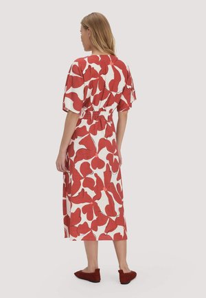 Frau mit blondem Haar, die ein weißes Midikleid mit großen roten Blattmustern und passenden roten flachen Schuhen trägt, steht und blickt nach hinten.