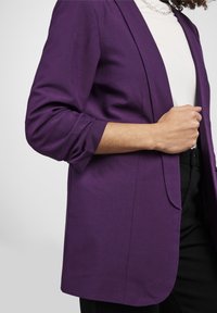 Personne portant un blazer violet à manches retroussées sur une chemise blanche, accessoirisée d'un collier chaîne en argent, et un pantalon noir.
