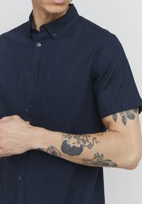 Granatowa koszula krótki rękaw zapinana na guziki, wykonana z lekkiego materiału, z kołnierzykiem typu wskazującego i z przodu na guziki. Widoczne tatuaże na przedramieniu.