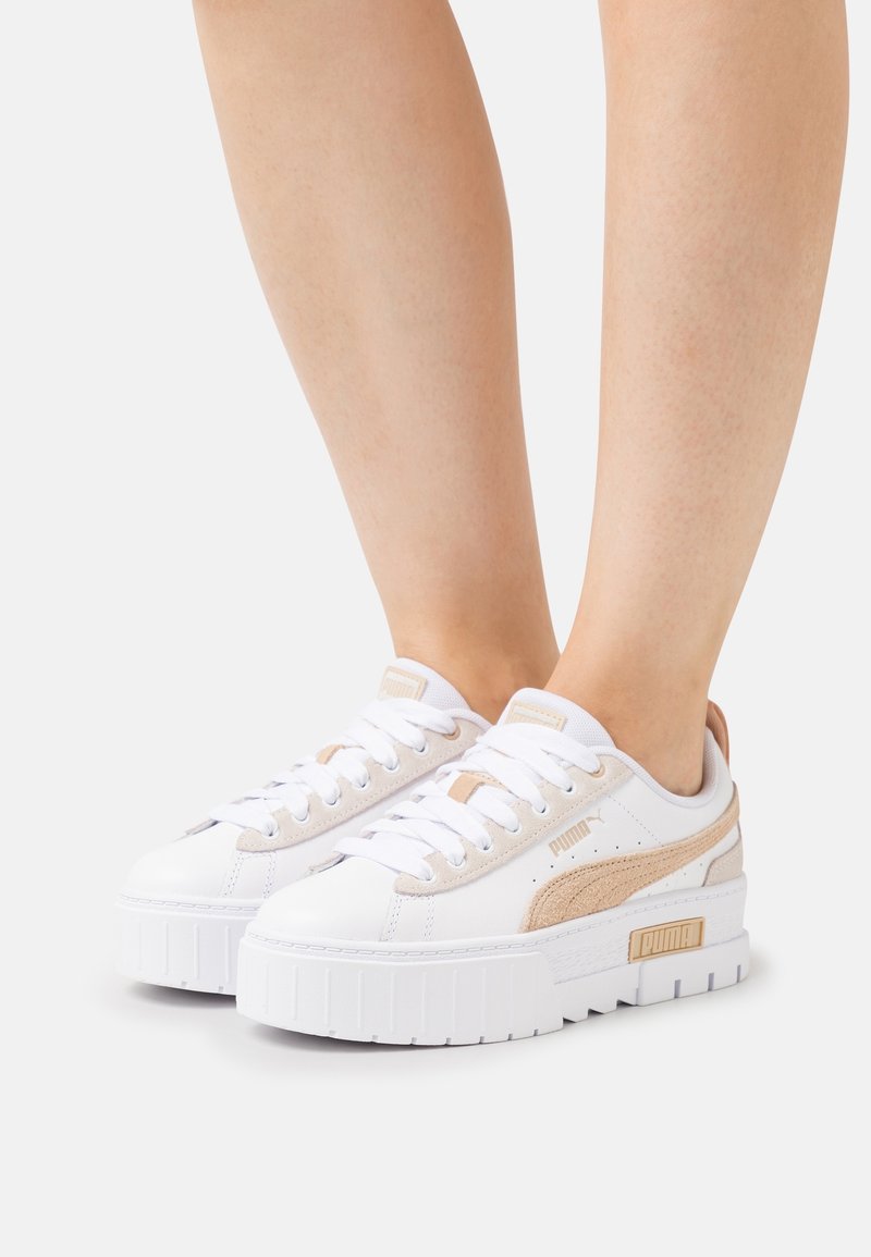 Jambes portant des baskets blanches Puma avec des bandes en daim beige et des semelles compensées épaisses et texturées sur un fond blanc uni.