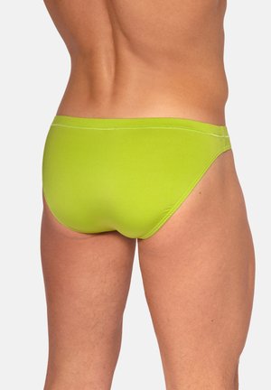 Mann in leuchtend grüner Badehose, Rückansicht, vor schlichtem hellgrauem Hintergrund.