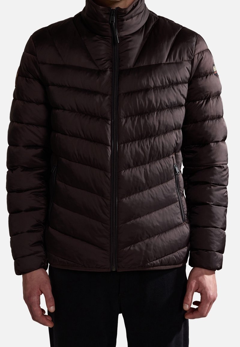 Mężczyzna ubrany w ciemnobrązową pikowaną kurtkę zapinaną na zamek z ukośnym przeszyciem i zamykanymi kieszeniami bocznymi.