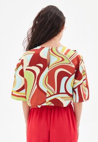 Blusa a maniche corte con motivi astratti multicolore in rosso, blu, verde e crema, con dettaglio a buco e bottone sul retro.