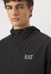 Μαύρο μπουφάν με φερμουάρ και ψηλό γιακά, διαθέτει μικρό λογότυπο EA7 στο αριστερό στήθος, κατασκευασμένο από λείο, ελαφρύ ύφασμα.