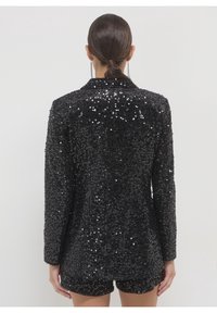 Femme aux cheveux foncés en chignon bas, portant un blazer noir à sequins à manches longues et un short assorti, vue de dos sur un fond uni.