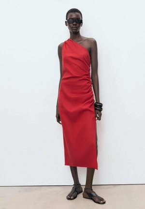 Robe rouge à une épaule avec des détails froncés sur le côté, longueur genoux, en tissu lisse. Portée avec des lunettes de soleil noires et des sandales.