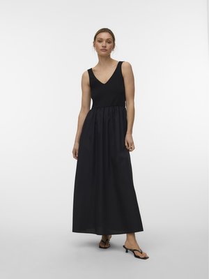 Kvinne iført en svart ermeløs maxikjole med V-hals og samlet midje, stående i svarte sandaler med hæl på ensfarget bakgrunn.