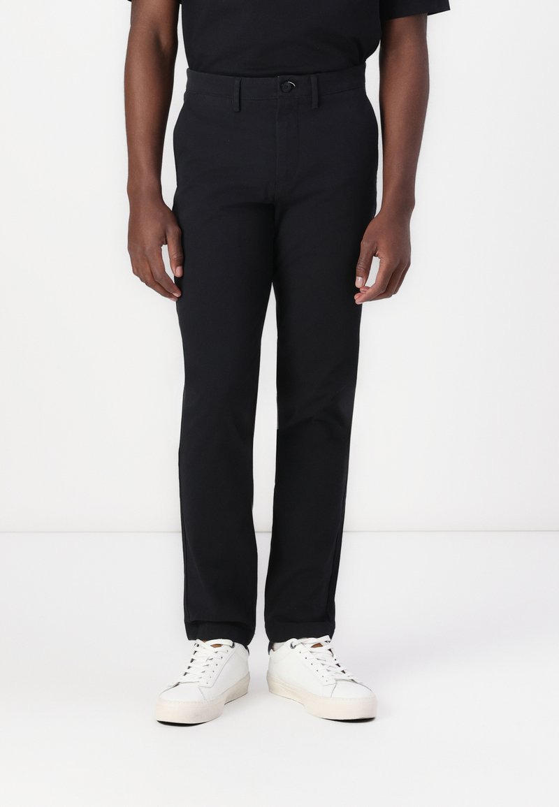 Black tailored trousers with a button closure, belt loops, and a slim fit design. Paired with white sneakers for a casual look.