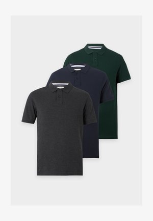 Homme portant un polo vert foncé, un jean bleu clair et des baskets marron, debout avec une main dans la poche devant un fond blanc.