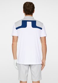 White polo shirt with a gray and navy blue pattern on the back, short sleeves, and a flat collar, paired with light gray shorts and a glove.