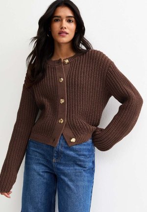 Donna che indossa un cardigan lavorato a maglia marrone con bottoni dorati e jeans azzurri, in piedi con una mano sui fianchi contro uno sfondo bianco.