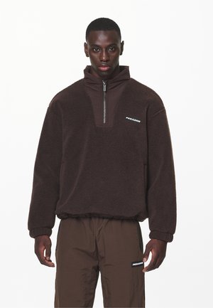 Brown fleece pullover with a high collar, half-zip front, and elastic cuffs. Features a branded logo on the chest. Lightweight, textured fabric.