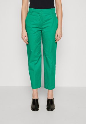 Pantalons en coton verts avec une coupe ajustée, dotés d'une taille mi-haute, de poches et de poignets repliés, associés à des chaussures noires à enfiler.