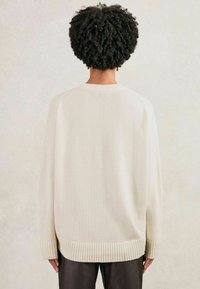 Persoană cu păr creț scurt, purtând un pulover oversized crem și pantaloni închiși la culoare, întoarsă cu fața la un fundal deschis simplu.