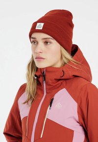 Rode en roze skijas met een hoge kraag, een blinde rits en waterdicht materiaal. Een kastanjebruine beanie met een logo patch maakt de look compleet.