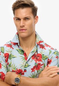 Camisa de botones de manga corta con un fondo azul claro, diseño floral rojo y hojas verdes. Presenta un cuello extendido y detalles en botones.