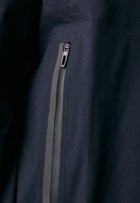 Veste marine dotée d'une fermeture éclair horizontale, d'une texture de tissu lisse et d'un contraste de bord noir le long de la ligne de la fermeture éclair pour une durabilité accrue.