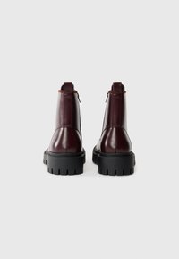 Botines de cuero burdeos con acabado mate, que presentan una suela de goma negra gruesa, cierre de cremallera y un acento en color marrón claro en el talón.