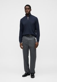 Μπλε ναυτικό μακρυμάνικο τοπ με φερμουάρ, γκρι παντελόνι με σχέδιο, μαύρη ζώνη και λαμπερές μαύρες παπούτσια. Το μοντέλο στέκεται μπροστά σε απλό λευκό φόντο.