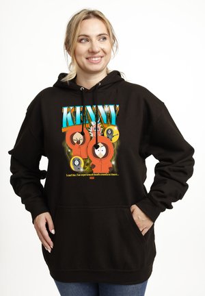 Uśmiechnięta kobieta w czarnej bluzie z kolorową grafiką Kenny’ego z South Park i tekstem o doświadczaniu śmierci niezliczoną ilość razy, ręce w kieszeniach.