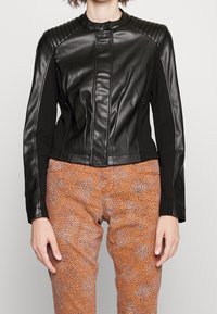 Schwarze Lederjacke mit gerippten Schulterakzenten, schmaler Passform und hohem Kragen, kombiniert mit gemusterten braunen Hosen.