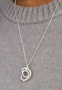 Silverhalsband med en abstrakt, sammanflätad design på en tunn kedja, uppvisad mot en ljusgrå stickad tröja.