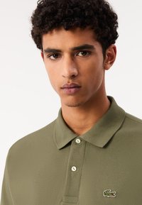 Khaki pikétröja i texturerat tyg, med krage, tre knappar och en grön krokodil-logotyp på vänster bröst.