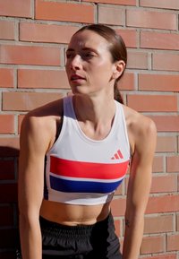 Biała sportowa crop top z czerwonymi i niebieskimi poziomymi paskami, z logo Adidas. Czarne szorty sportowe, na tle ceglanej ściany.