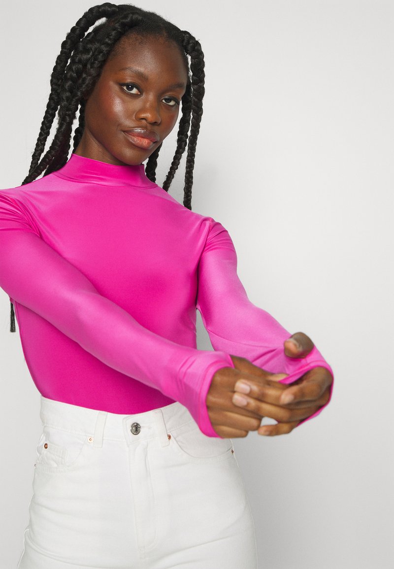 Haut ajusté à manches longues de couleur rose vif, en matériau lisse et extensible, avec un col haut et des ouvertures pour les pouces. Porté avec un jean blanc taille haute.
